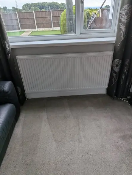 A newly fitted white panel radiator sits under a window in a carpeted room beside a dark leather sofa, with a garden and fence visible outside