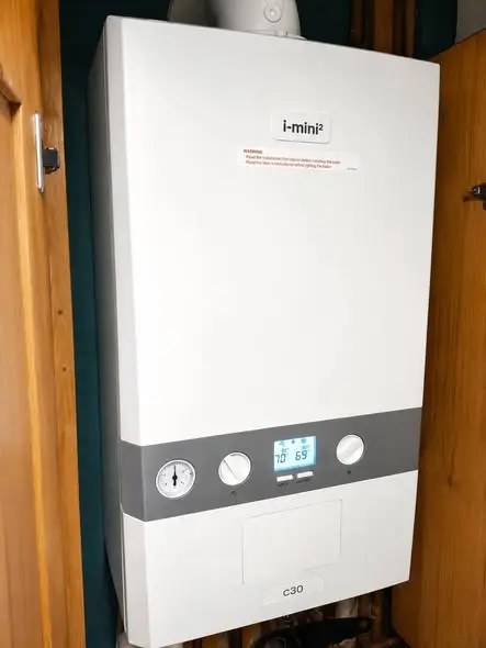 A wall-mounted white combi boiler with a grey control panel, pressure gauge, digital display and the model badge 'i-mini²' and 'C30', fitted in a tidy cupboard with wooden doors