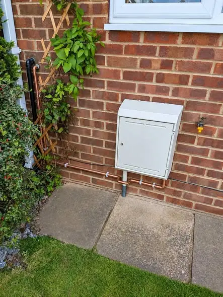 A white metal box, likely a gas meter enclosure, mounted on a red brick wall beside a window, with copper pipework running along the wall and an outside tap, above a paved path and lawn