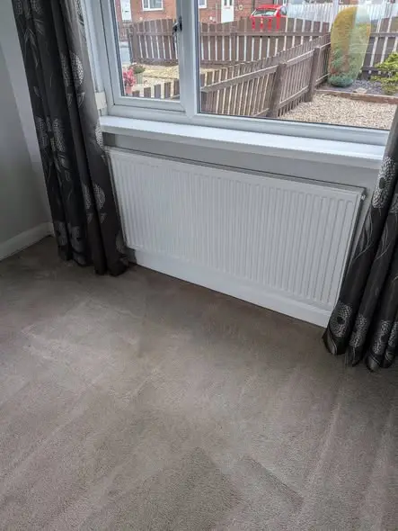A new white panel radiator is fitted beneath a window in a carpeted room with dark patterned curtains, with a garden fence visible outside