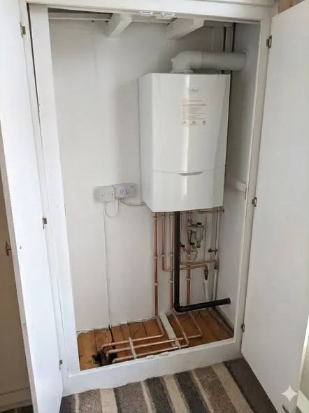 A wall-mounted white combi boiler, labelled Ideal, installed in a white-painted cupboard alcove with copper pipework, a flue running out the top, and a double socket beside it