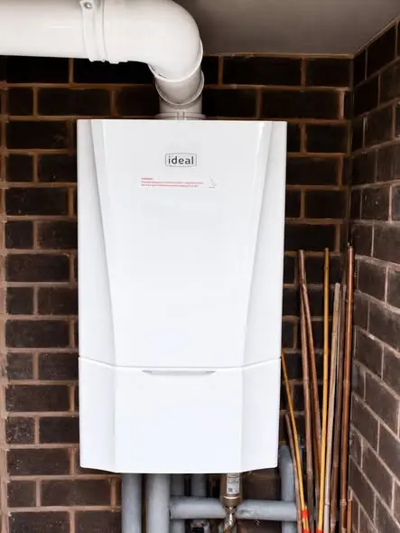 A newly installed white wall-mounted Ideal boiler with a flue pipe running out at the top, set against a dark brick wall with copper pipework and tails below it